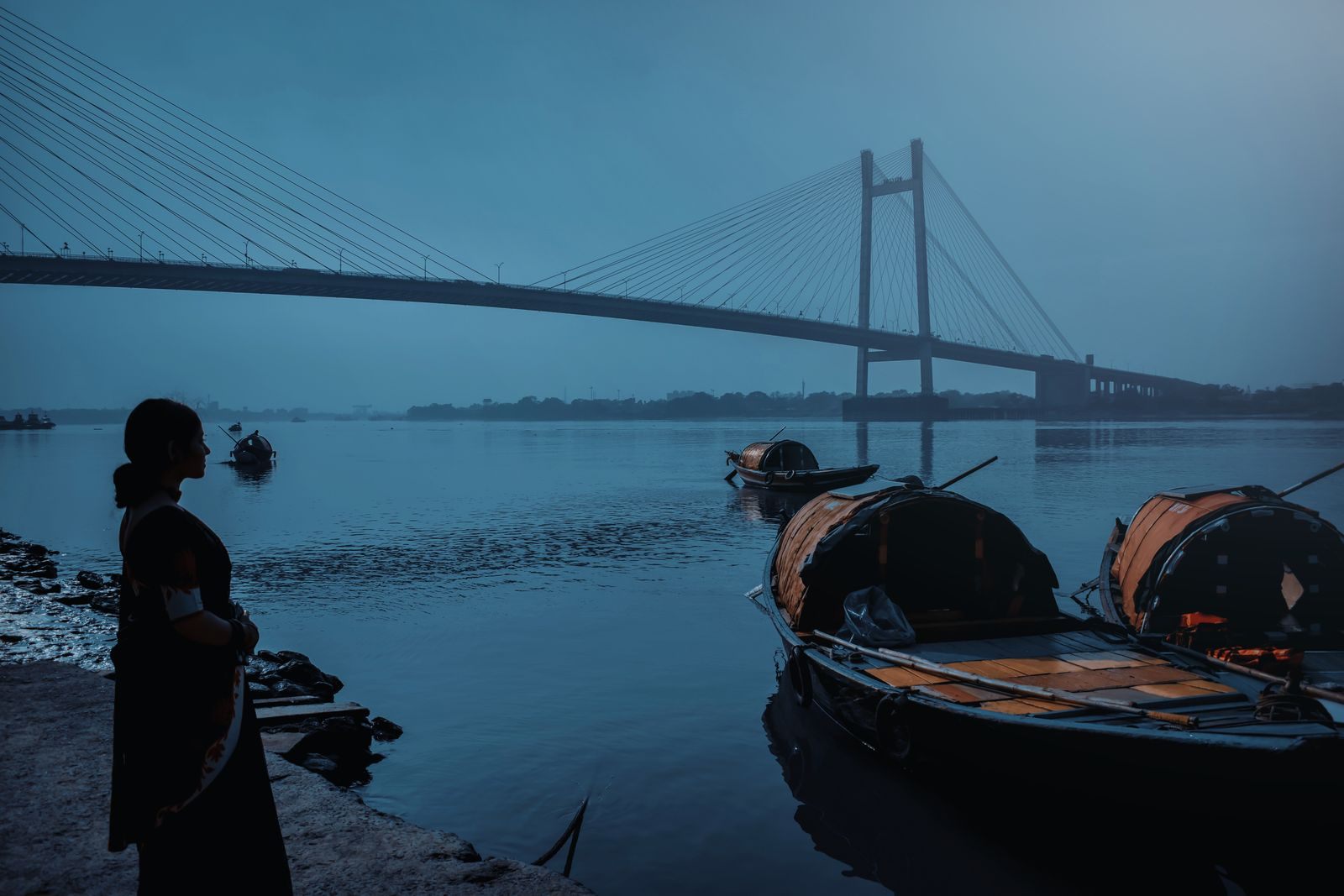 Moored wooden boats on the Hooghly at dawn with a cable-stayed bridge behind in Kolkata